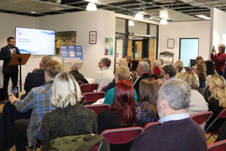 Zoomed out shot of VODA AGM 2024. Lots of people listening to speaker in the back of the image next to a large screen,