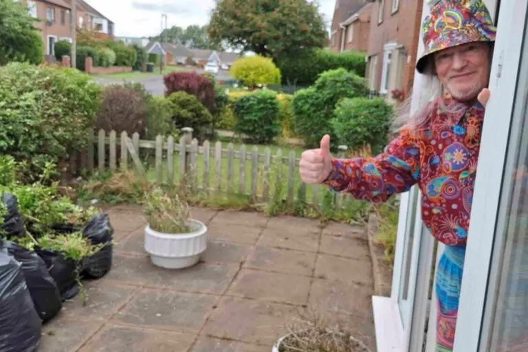 Tidy garden with happy person leaning out of door with their thumbs up. They are wearing red and have a brown hat on