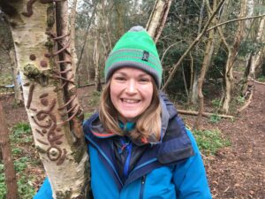 Alice wearing blue coat and green hat, leaning against tree