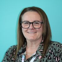 Vicky has dark hair, glasses and is wearing a green animal print top. She is smiling at the camera, in front of a blue/green wall.