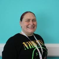 Leah is standing in front of a turquoise wall. Wearing a black tee with writing on the front. Leah has red hair in a pony tail and is smiling.