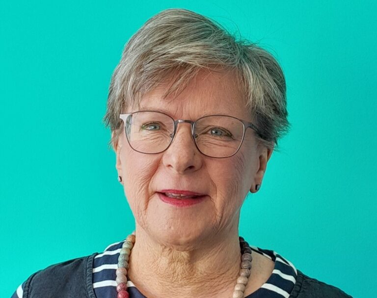 Penny has short light hair and glasses. She is wearing a dark top and necklace in front of a blue background.