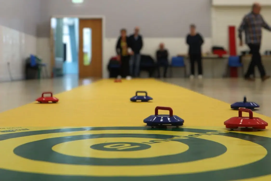 Yellow and green indoor kurling target in foreground with kurlers standing in the distance.