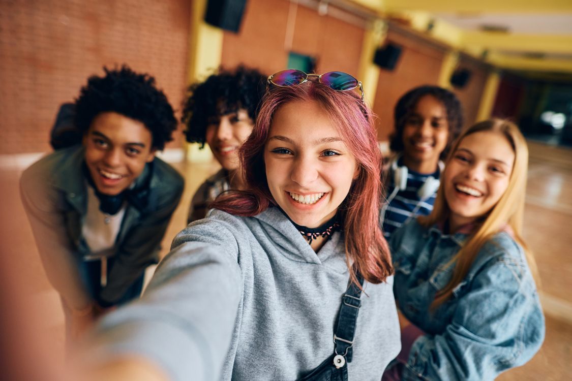 Group of young people taking a selfie. The person taking the pic is front centre, with pink hair, sunglasses on her head and a grey sweatshirt.