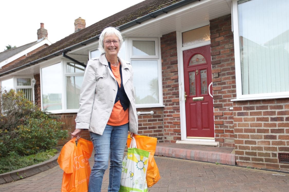 Volunteer outside of house carrying bright coloured shopping bags.