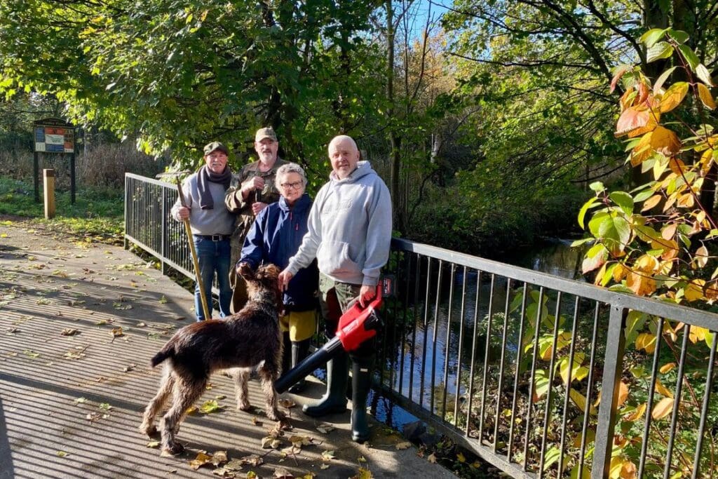 4 people and a large dog standing on a bridge. You can see lots of trees and shrubs behind them, as well as a small river. The people are holding tools and a leaf blower.