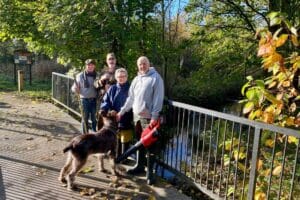 4 people and a large dog standing on a bridge. You can see lots of trees and shrubs behind them, as well as a small river. The people are holding tools and a leaf blower.