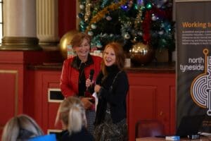 Two people in front of audience, Christmas tree is behind them. One is wearing red, the other black., They are both smiling.