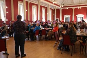 Shows the back of a presenter at MHA event, in front of them is a large group of people sat at tables. The room has red walls and big windows