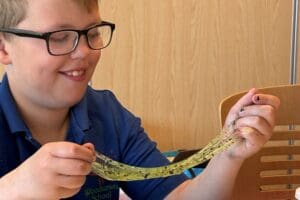 Smiling young boy wearing glasses and blue shirt stretching slime between his hands.