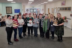 Group of women wearing festive jumpers and headwear. Two people are holding cake stands full of sweet treats.