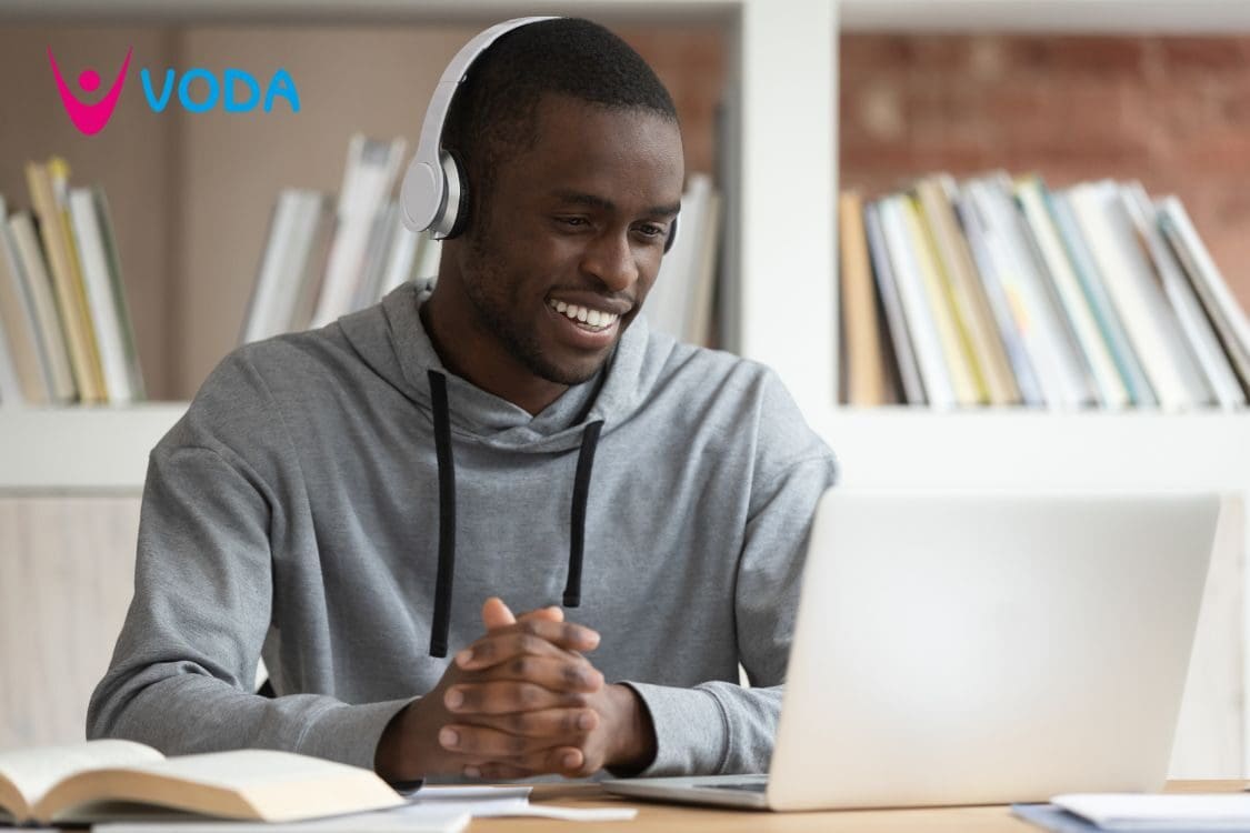 A smiling man in a grey hoody with headphones and a laptop. There are books on the shelves behind him.