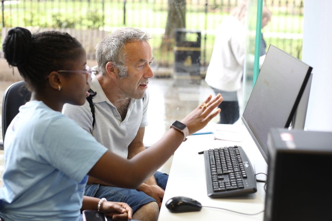 A man and a woman sitting in an office, looking at a computer screen together. They are dressed in professional clothing and appear to be engaged in work-related activities. The setting includes various office equipment and furniture.