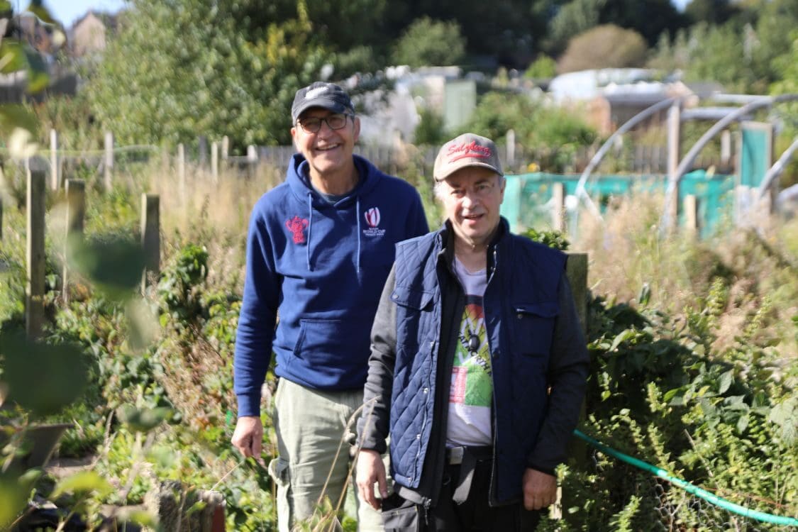 A couple of men standing in a garden with greenery, including plants and trees, while smiling on the grass. They are wearing casual mostly blue clothing.