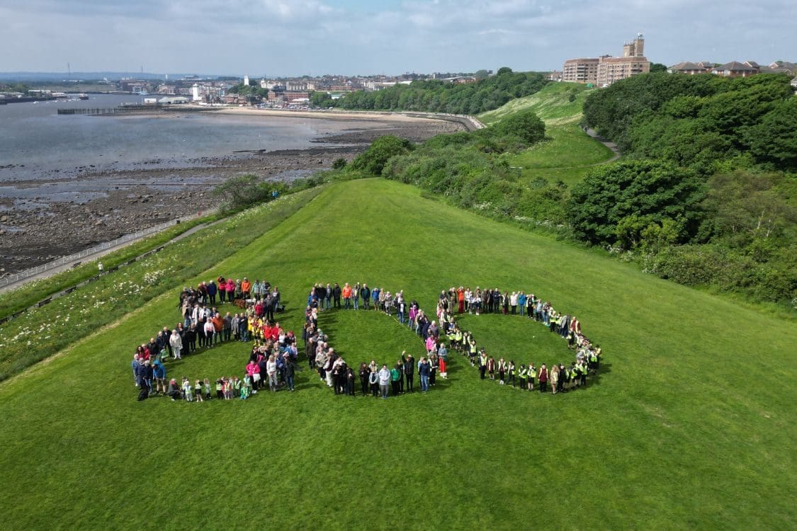 Large grassed area with North Shields in the background and the river tyne on the left. A large group of people are spelling out 800 on the grass.