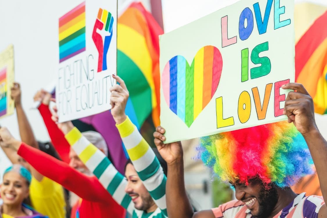 A diverse group of people smiling and holding colorful signs that say "LOVE IS LOVE." They are wearing vibrant clothing, and the scene reflects a joyful festival atmosphere with rainbow colors.