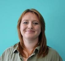 A head shot of Ellen with shoulder length red/blonde hair, wearing a khaki shirt Infront of a turquoise wall