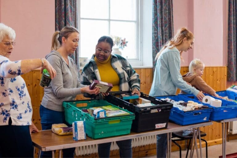 A group of people sorting food into boxes. There is a window behind them and the boxes are various coloured plastic stacking trays. There is a small child helping.