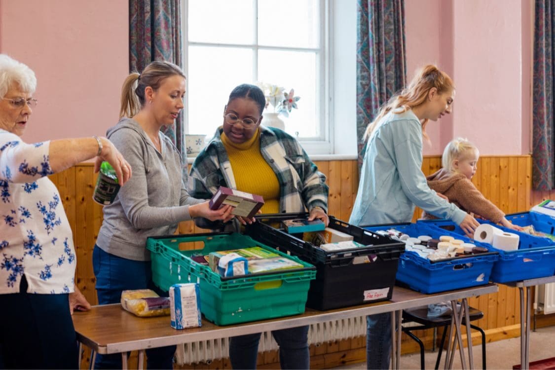 A group of people sorting food into boxes. There is a window behind them and the boxes are various coloured plastic stacking trays. There is a small child helping.