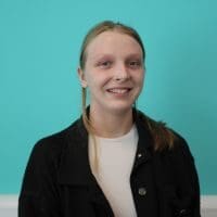 Headshot of Rosie in front of of a turquoise wall. Rosie is wearing a black jacket over a white tee and has fair hair which is tied back and over one shoulder. Rose is smiling