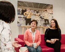 Two people sat on a pink sofe, there is a large picture behind them. One is wearing peach and the other is wearing black. Both are holding cups and are engaged in conversation with someone in the foreground with their back to the camera.