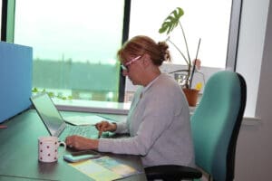 Side profile image of person using a laptop, sat on green chair. There is a window behind them and you can see rooftops. 
