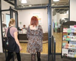 Two people walking into an office, In the background there is a reception desk with someone sat behind it. There is a leaflet stand to the righ of the image.