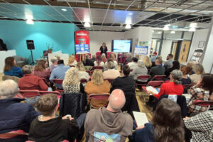 Large group of people sat facing a speaker at the back of the image.