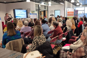 Large group of people sat facing a speaker at the left side of the image.