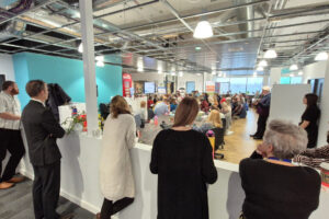 Behind the scenes shot of VODA AGM. Staff are at the side of the event, image captured from behind. The staff team is watching the agm and looking out towards a large crowd of seated people.