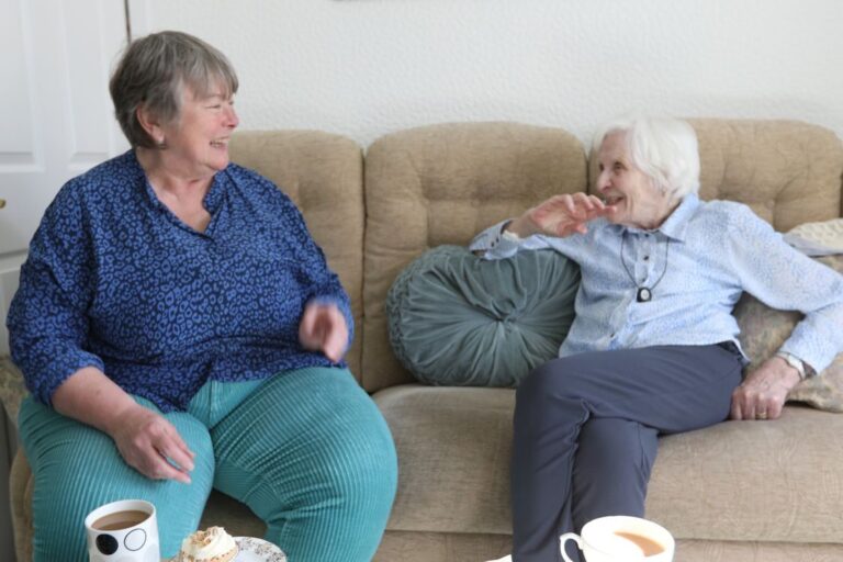 Two women sat on a brown sofa. Both are wearing blue. They are smiling and chatting over a cuppa and some cake.
