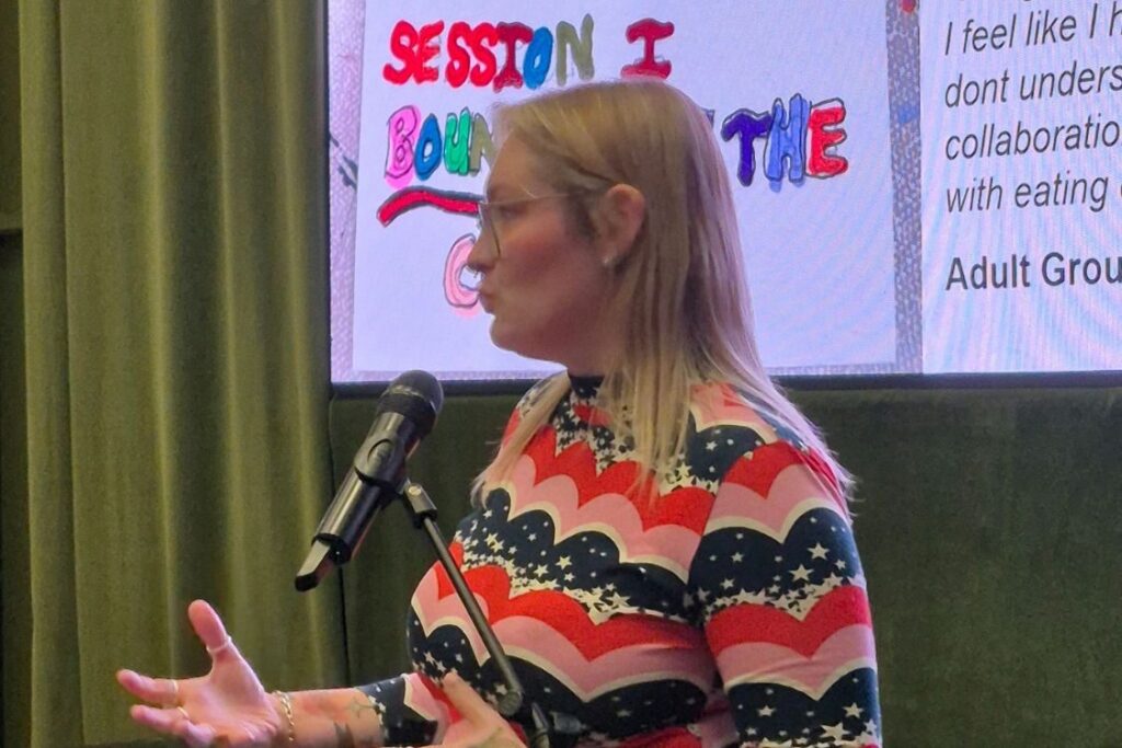 Presenter with blonde hair, glasses and stripey top ion front of a screen