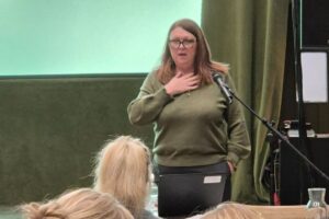 Presenter with green top, glasses and brown hair talking to audience