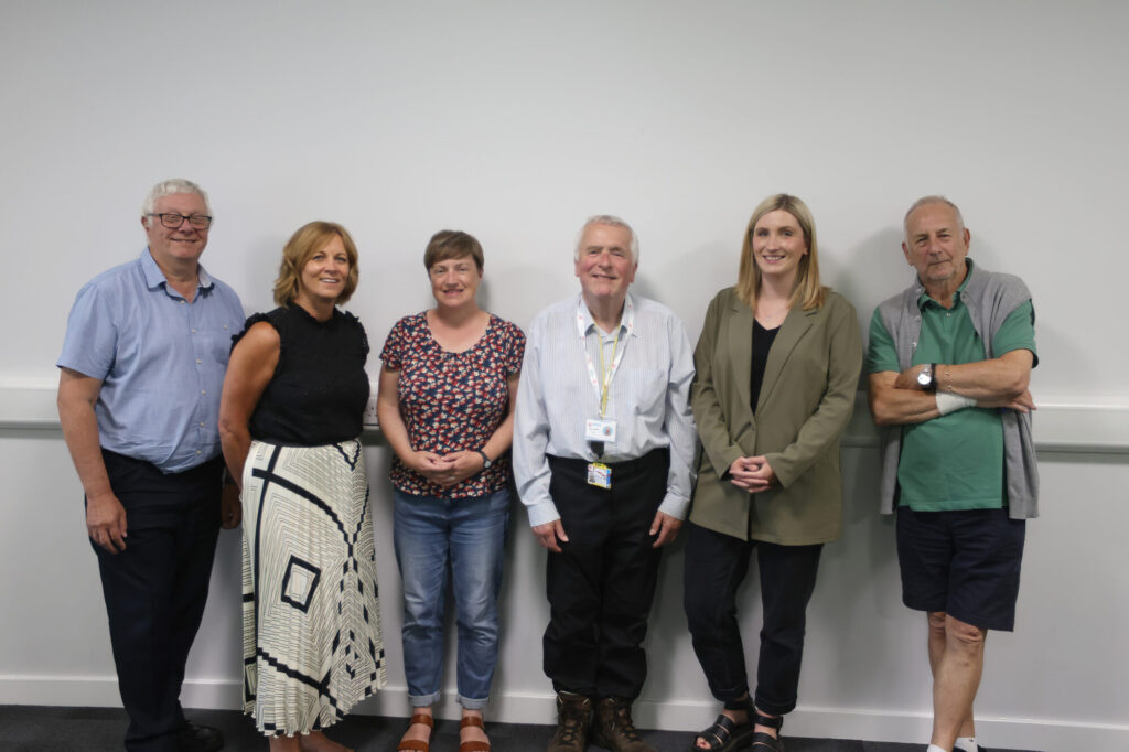 Six people lined up in front of a white wall. They are wearing a variety of clothes and are smiling at the camera.