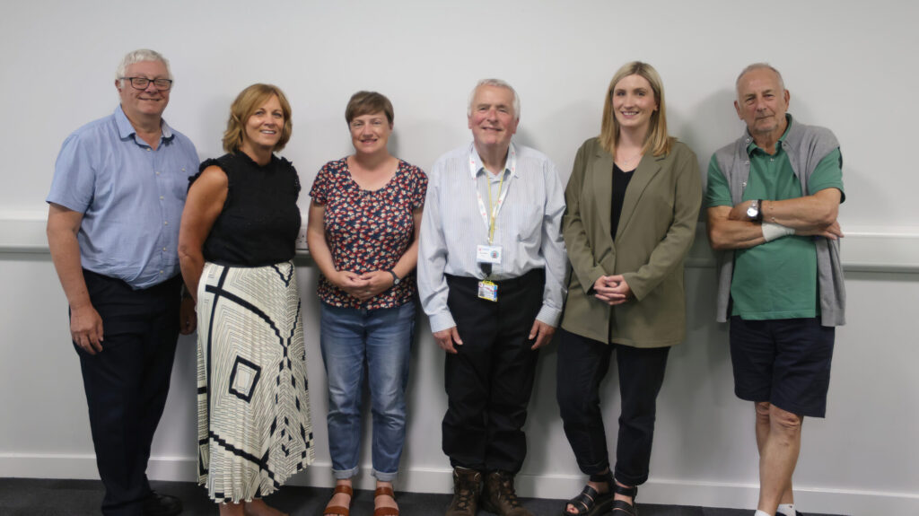 Six people lined up in front of a white wall. They are wearing a variety of clothes and are smiling at the camera.