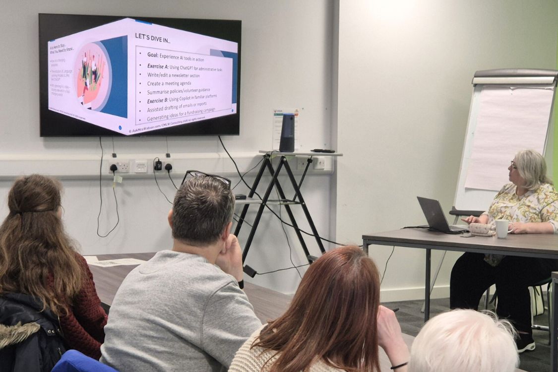 A group of people in a workshop environment facing away from the camera looking at a presentation on screen about AI.