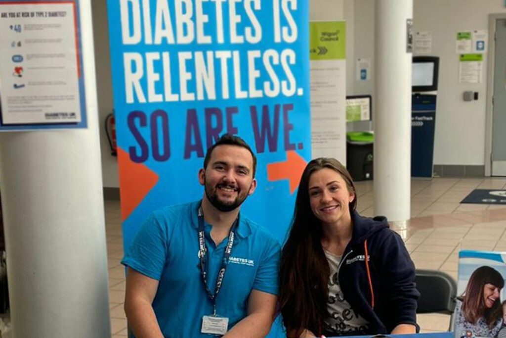 Two people sit at an information stand inside a public building, positioned in front of a large blue banner that reads “DIABETES IS RELENTLESS. SO ARE WE.” in bold white lettering with orange accents. Both individuals wear blue Diabetes UK-branded clothing and lanyards. The setting appears to be an indoor venue with tiled floors, white pillars, and informational posters in the background.