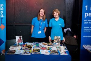 Two people stand behind a table covered with a blue cloth and filled with a wide range of diabetes-related leaflets, brochures, and booklets. Both are wearing blue Diabetes UK polo shirts. The table display includes printed materials with photos and headings, arranged in neat piles. A dark backdrop and a vertical banner frame the stand, indicating a health information or outreach event.