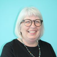 A headshot of Lisa with white hair in a bob. Lisa is wearing glasses and smiling at the camera. Wearing a dark top with a blue/green wall behind her