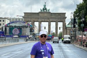 Man wearing white hat and purple volunteer shirt in front of brandburgh gate, berlin