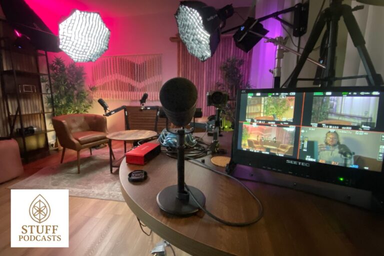 Pink lit podcast studio with chairs, mic's and video screens. Stuff Podcasts logo