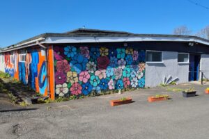 Exterior of a low, single-storey building on a sunny day, featuring a large, colourful mural of flowers painted across a brick wall. The artwork includes bright blues, pinks, yellows and purples. Part of the building is painted white with a blue door and small windows. Several rectangular planters sit on the paved ground in front of the building, with some greenery starting to grow.