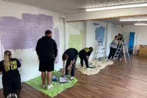 Indoor space where a group of people are painting a large wall. The room has wooden flooring, white brick walls and strip lighting on the ceiling. Several people are using rollers and brushes, with paint trays and protective sheets on the floor. Different sections of the wall are being painted in light colours such as purple, green and blue, and a step ladder is set up on the right-hand side.