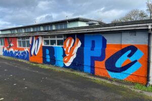 Exterior of a single-storey building with a large, colourful mural painted across a long brick wall. The mural features bold lettering in blue, orange and white, alongside a bird and wave design. The building has a row of windows near the roofline, a paved area in front, and a cloudy sky overhead.