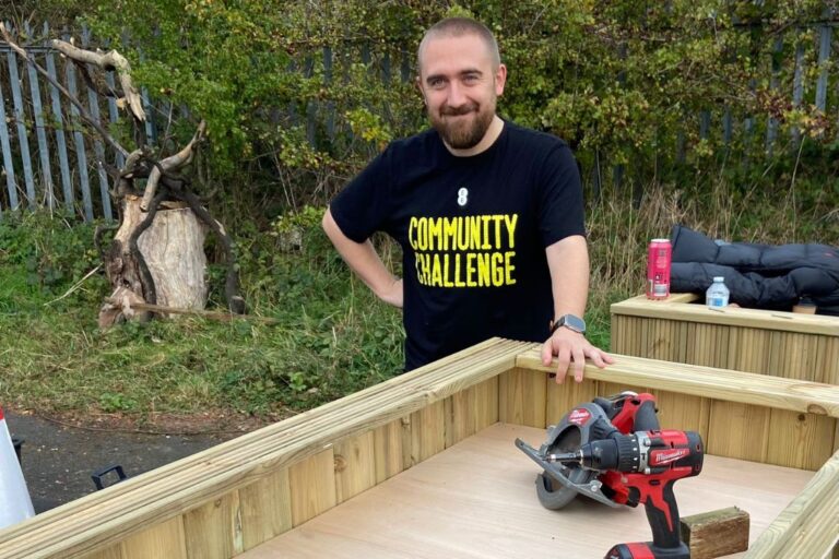 Outdoor scene showing a person standing beside a partially constructed wooden planter or raised bed. Tools, including a red power drill and circular saw, are placed on the wooden surface. In the background there is greenery, a fence and some outdoor items like bottles and a jacket. The person is wearing a black T-shirt with the words “COMMUNITY CHALLENGE” printed on it.
