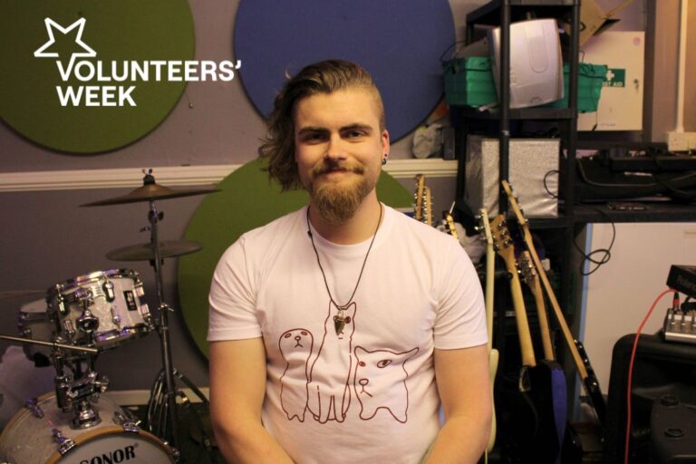 Young man with longish hair and beard wearing white t-shirt sits in front of music equipment including drums and guitars