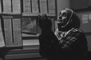 Black-and-white photograph of a person with hands raised beneath sheets displaying dates and numbers, featured in the Asylum: A Long and Painful Process photography exhibition.