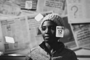 Black-and-white portrait of a person wearing a knitted hat, surrounded by hanging date cards and newspapers, displayed as part of the Asylum: A Long and Painful Process photography exhibition.