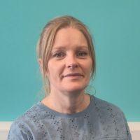 Picture of Joanne, facing the camera, she has blonde hair tied back and a light blue top.