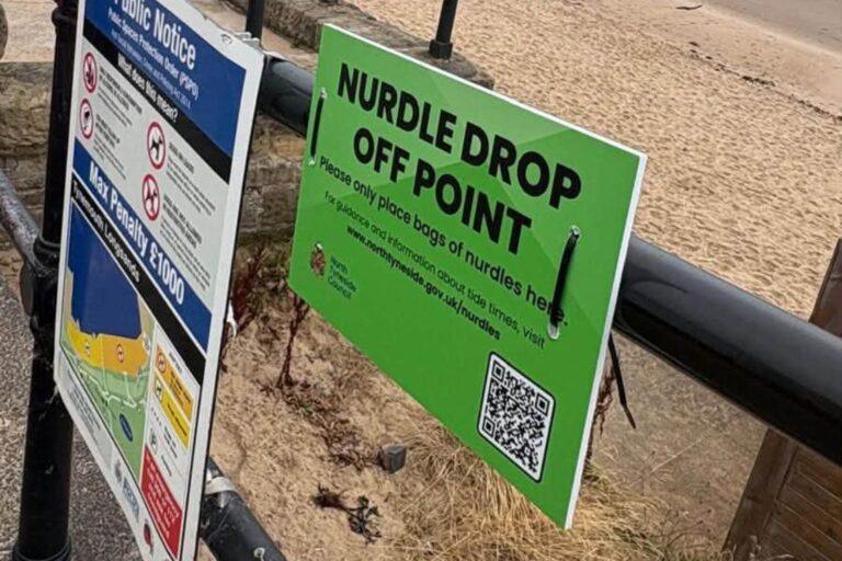 Picture of a green sign on a black railing which says 'nurdle drop off point' there is a beach in the background and a public notice sign to the left